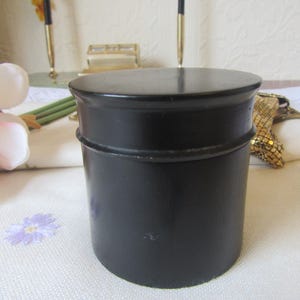 May include: A black, cylindrical container with a matching lid. The container has a simple, classic design, with a slightly rounded top and bottom. The surface appears smooth and matte. The container is likely used for storage or display.