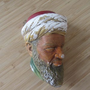 Op de afbeelding: Gedetailleerde sculptuur van een mannenhoofd, met een witte en gouden tulband met rode accenten. Het gezicht heeft een verweerde uitstraling met een lange, grijze baard en een groene sjaal. De sculptuur staat op een lichtbruin houten oppervlak.