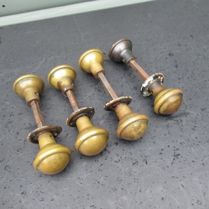 Peut inclure: Quatre boutons de porte anciens en laiton avec une patine vieillie. Chaque bouton a une forme arrondie avec une base décorative et une longue tige élancée. Les boutons sont disposés sur une surface sombre et texturée.