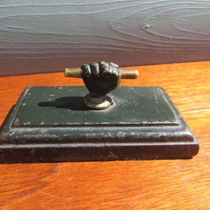 May include: An antique green and black paperweight with a hand-shaped handle. The handle is dark with a brass rod. The paperweight is rectangular and sits on a wooden surface.