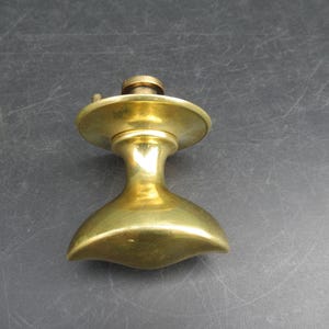 May include: A polished brass door knob featuring a wide, curved, horizontal handle that tapers at both ends. The knob includes a circular backplate and a central cylindrical mounting component with a small screw visible on the top surface.