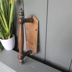 May include: A vintage-style door handle featuring a vertical, cylindrical grip with decorative finials at the top and bottom. The handle is attached to a rectangular brass backplate with rounded corners and four visible screw holes for mounting.