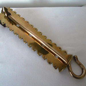 May include: A brass doorstop with a decorative scalloped edge and a hook on one end. The doorstop is designed to hold a door open.