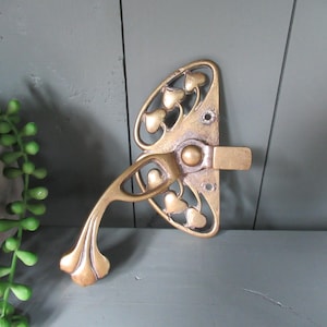 May include: An antique brass door latch with a decorative design. The latch features an oval backplate with heart-shaped cutouts and a curved handle. The metal has a warm, aged patina, suggesting its vintage origin. The latch is mounted on a gray surface.