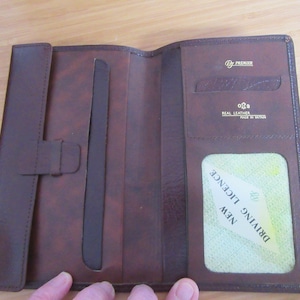 Può includere: Un portafoglio in pelle marrone scuro con chiusura a patta verticale, scomparti multipli per carte e una finestra in plastica trasparente contenente una carta con il testo NEW DRIVING LICENCE. L'interno reca il marchio Premier e Real Leather Made in Britain.