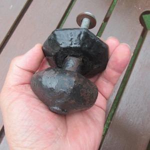 May include: A black, rustic door knob with a textured finish. The knob features a faceted top and a rounded base, joined by a metal stem. A washer and bolt are visible at the top. The knob is held in a hand.