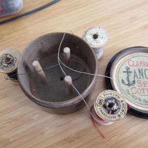 May include: A vintage sewing kit with wooden spools of thread, a brown container with wooden pegs, and a lid with "Clark & Co Anchor Sewing Cottons" text. The kit is on a light brown wooden surface.