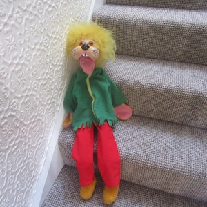 May include: A vintage handmade puppet with a lion-like face and a long, pink tongue. It wears a green jacket with a yellow zipper, red pants, and yellow shoes. The puppet is positioned on a staircase with gray carpet.