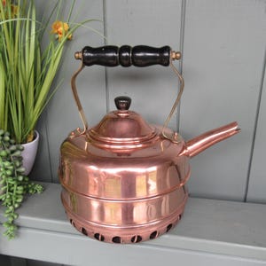 May include: A polished copper kettle featuring a cylindrical body with a tiered design, a wide angled spout, and a black wooden handle. The lid is topped with a small black knob, and the base includes a series of circular ventilation holes.