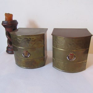 Op de afbeelding: Twee antieke messing dozen met een afgeronde bovenkant en een decoratief embleem aan de voorkant. De dozen staan op een wit oppervlak.