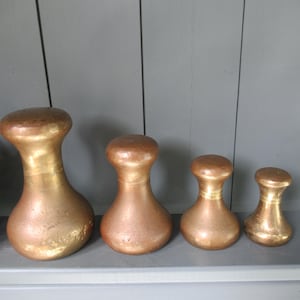 May include: Four decorative objects in metallic gold. Each has a rounded top and a bulbous base, similar to bowling pins. They are arranged in a row, with the largest on the left and smallest on the right. The tallest is approximately 20 cm high.