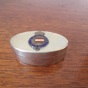 May include: A silver oval-shaped trinket box with a blue and gold crest featuring a flag with the letters "YB" on it.