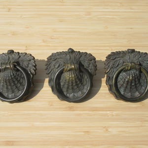 Op de afbeelding: Drie antieke bronzen deurkloppers met een decoratief bladerenontwerp en een ringhandvat. De kloppers zijn in een rij gerangschikt op een lichtbruin houten oppervlak. De hardware heeft een verweerde uitstraling.