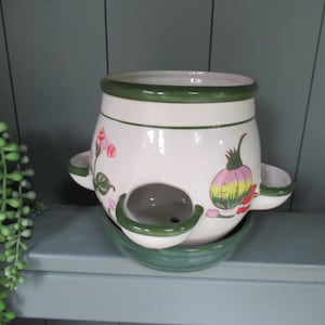 May include: A white ceramic herb pot with a green rim and base, featuring pink floral and vegetable illustrations. The pot has three side compartments and a central opening, ideal for growing herbs. The pot measures approximately 18 cm in height.