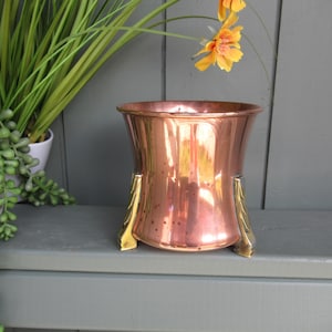 Può includere: Un portavaso in rame lucido con bordo svasato e tre piedini in metallo dorato decorati. Il contenitore presenta una superficie liscia e riflettente con un'ampia apertura, sostenuto da gambe decorative che sollevano la base.