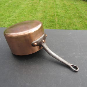 May include: A copper saucepan with a long, riveted iron handle. The pot has a rounded base and a lid. The copper has a warm, reddish-brown hue, and the handle is a dark gray color. The saucepan is likely used for cooking.