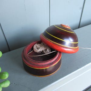 Puede incluir: Un costurero de madera vintage en marrón oscuro, con rayas rojas y amarillas. La tapa está abierta, revelando un carrete de hilo. El costurero descansa sobre una superficie gris, sugiriendo un tema de artesanía o costura.