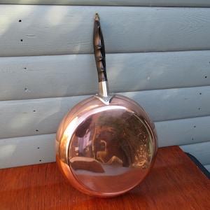 May include: A copper-colored, round-bottomed pan with a long, dark wooden handle. The pan has a shiny, reflective surface. The handle is attached with a silver-colored metal piece. The pan is sitting on a wooden surface.