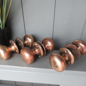 Könnte beinhalten: Ein Set aus drei polierten Türknäufen aus Kupfer, jeweils mit einem glatten, abgerundeten Griff und einer runden Montageplatte mit sichtbaren Schraublöchern. Die metallische Oberfläche reflektiert das Licht in warmen Tönen.