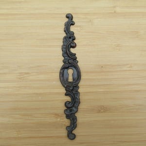 Könnte beinhalten: Ein dunkelbraunes Metall-Schlüsselschild mit einem länglichen, vertikalen Design, verziert mit geschwungenen Ornamenten und einer zentralen Schlüssellochöffnung. Der Beschlag verfügt über zwei kleine Schraublöcher an den Enden.