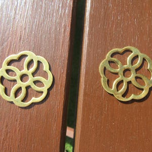 以下が含まれることがあります： 真鍮色の、透かし彫りの花型の装飾金具が2つ。金具は茶色の木製の表面に配置されています。花の模様は、中央の円と6つの湾曲した花びらがあります。