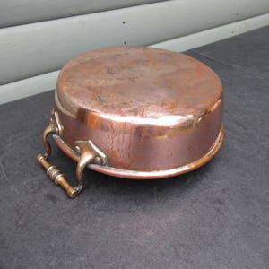 以下が含まれることがあります： 丸みを帯びた底と平らな上面を持つ銅製の調理鍋。鍋は、片側にブロンズのハンドルが付いた、変色した、古くなった外観をしています。鍋は暗い表面に置かれており、ヴィンテージまたはアンティークであることを示唆しています。