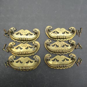 Peut inclure: Six poignées de tiroir en laiton avec des plaques arrière ajourées et ornées, accompagnées de poignées incurvées assorties. Chaque poignée présente un élément décoratif central et deux points de fixation. Des vis métalliques sont incluses.