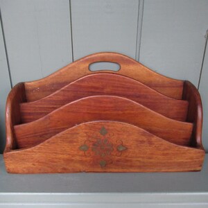 May include: A wooden letter rack with three sections and a handle. The rack is made of reddish-brown wood and has a decorative brass-coloured design on the front. The design is a floral pattern.