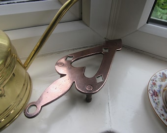 May include: A copper-colored trivet with a heart-shaped design and a handle. The trivet has a decorative pattern with square cutouts and is resting on a white surface. A brass watering can is partially visible in the background.