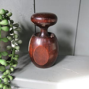 Op de afbeelding: Een donkerbruine houten flesstopper met een afgeronde bovenkant en een bolvormige basis. De stopper heeft een cirkelvormig accent aan de voorkant en wordt weergegeven op een wit oppervlak. Een groene plant is zichtbaar aan de linkerkant van de afbeelding.