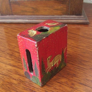 May include: A small, red wooden box with a painted design of birds and deer. The box has a slot on the side and is decorated with green and gold accents.