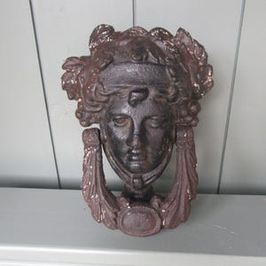 Può includere: Un battente per porta in ghisa raffigurante un volto femminile classico con capelli ornati e una cornice decorativa. Il battente è marrone scuro con dettagli dorati. È montato su una superficie grigio chiaro.