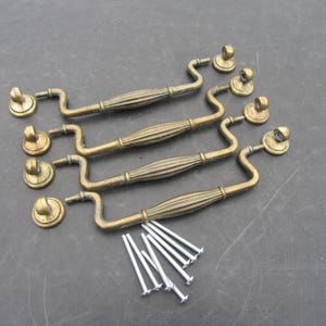 Op de afbeelding: Set van vier antieke messing lade-trekkers met bijpassende achterplaten en schroeven. De trekkers hebben een geribbeld middenstuk en gebogen handgrepen. De hardware heeft een warme, verouderde messingkleur, ideaal voor restauratie.