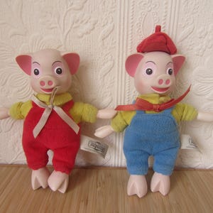 Può includere: Due peluche a forma di maialino. Uno indossa una tuta rossa e una camicia gialla, l'altro indossa una tuta blu e una camicia gialla con un berretto rosso. Entrambi hanno orecchie, musi e piedi rosa.