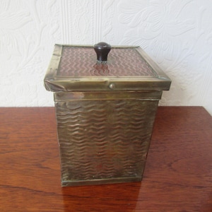May include: A vintage brass box with a textured wave pattern on the sides. The box has a hinged lid with a dark brown knob and a patterned glass insert. The box is rectangular and sits on a wooden surface.