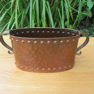 May include: An oval copper planter with two handles. The planter has a hammered finish and is decorated with small rivets around the rim.