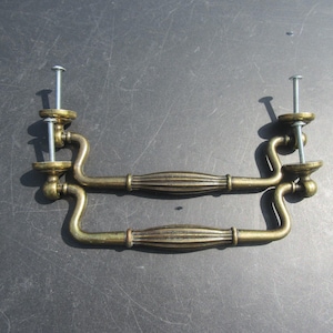 May include: Two antique brass drawer pulls with decorative ribbed centre sections and curved handles. Each pull has two mounting posts with screws. The hardware is a warm, aged brass colour, suggesting vintage or retro style.