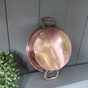 Puede incluir: Una olla redonda de color cobre con dos asas. La olla tiene una superficie brillante y reflectante y un borde ligeramente curvado. Las asas son de un color metálico más claro. La olla es probablemente vintage o antigua.