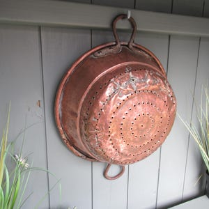 May include: A weathered copper colander with two handles hangs on a grey wooden wall. The colander features a series of small holes in a circular pattern. The copper has a worn appearance, suggesting age. Decorative kitchenware.