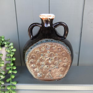 May include: A dark brown ceramic vase with two handles and a decorative, textured front. The vase has a light blue band near the top and a white rim. The front features a pattern of raised circles and a triangular symbol.