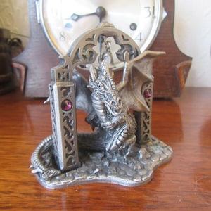 Könnte beinhalten: Eine Zinnfigur eines Drachen, der aus einem dekorativen Torbogen mit rosafarbenen Edelsteinen kommt. Der Drache hat ausgebreitete Flügel und ist mit Schuppen und Krallen verziert.