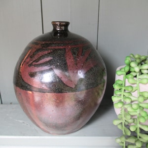 Può includere: Un vaso in ceramica fatto a mano, con corpo arrotondato e collo stretto. Il vaso presenta una parte superiore marrone scuro con disegni astratti rossi e neri. La metà inferiore sfuma in una tonalità marrone rossiccio. A destra, una piccola fioriera rosa chiaro con piante verdi pendenti.