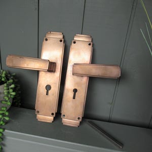Op de afbeelding: Een paar rechthoekige koperkleurige deurklinken met bijpassende schilden, elk voorzien van een centraal sleutelgat, getrapte randen en een horizontale hendel. De metalen oppervlakken hebben een gladde, metallic afwerking met subtiele kleurnuances.