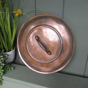 May include: A round copper lid with a polished metallic finish and a central handle. The lid features a circular recessed design and a smooth, slightly weathered surface texture, showcasing the natural reddish-brown tones of the copper material.