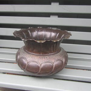 May include: A bronze-coloured decorative vase with a ruffled rim and a patterned lower section. The vase sits on a light grey bench. The vase's design features a textured pattern and a series of rounded shapes.