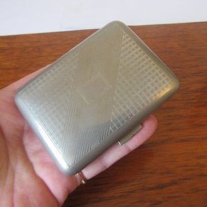 Puede incluir: Un estuche de cigarrillos de plata con un patrón geométrico y una tapa con bisagras.
