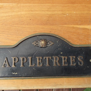 Op de afbeelding: Zwart metalen bord met de tekst "APPLETREES" in gouden letters. Het bord heeft een decoratieve bovenkant met een bloemenmotief en ligt op een houten oppervlak.