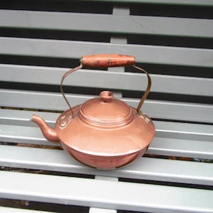 May include: A copper kettle with a wooden handle and a curved spout. The kettle has a lid and a handle that arches over the top. The kettle is sitting on a grey bench.