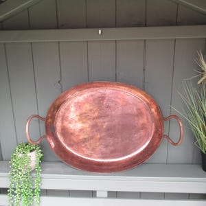 May include: Oval copper tray with two curved handles. The tray has a rich, reddish-brown colour and a slightly aged appearance. The tray is displayed on a grey bench with a green background and decorative plants.