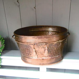 May include: A copper-coloured metal planter with a hammered texture. The oval container has a rope detail with tassels. It has two handles and sits on a white shelf. A green plant is visible on the left.